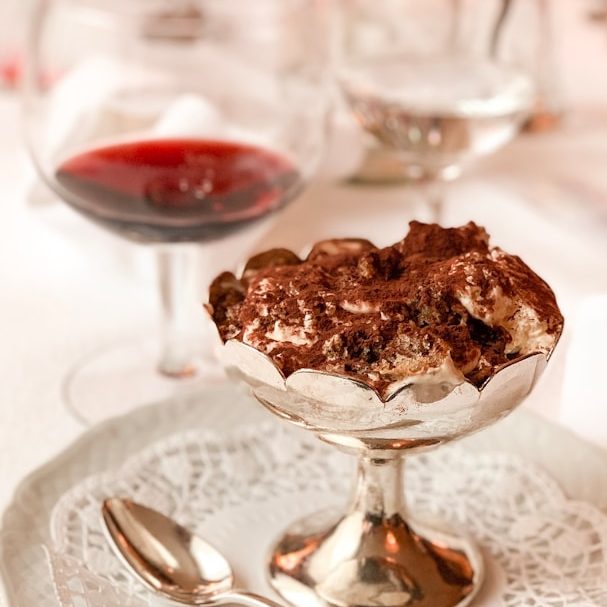 Schokoladendessert in einer silbernen Schale mit einem Weinglas im Hintergrund.
