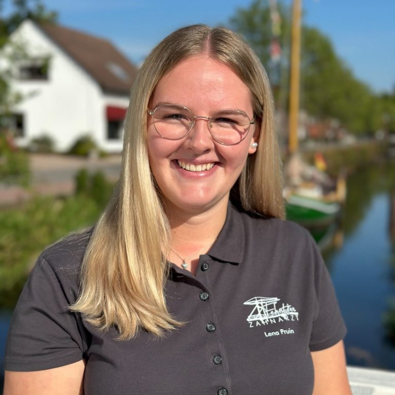 Junge Frau mit langen Haaren, Brille und grauem Hemd, lächelt vor einer Wasserlandschaft.