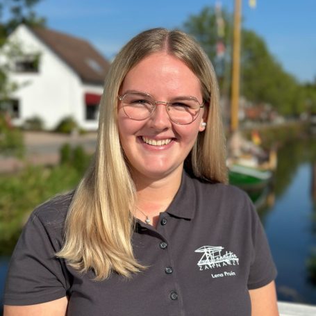 Lächelnde Person mit blonden Haaren in einem grauen Polohemd vor einem malerischen Kanal.