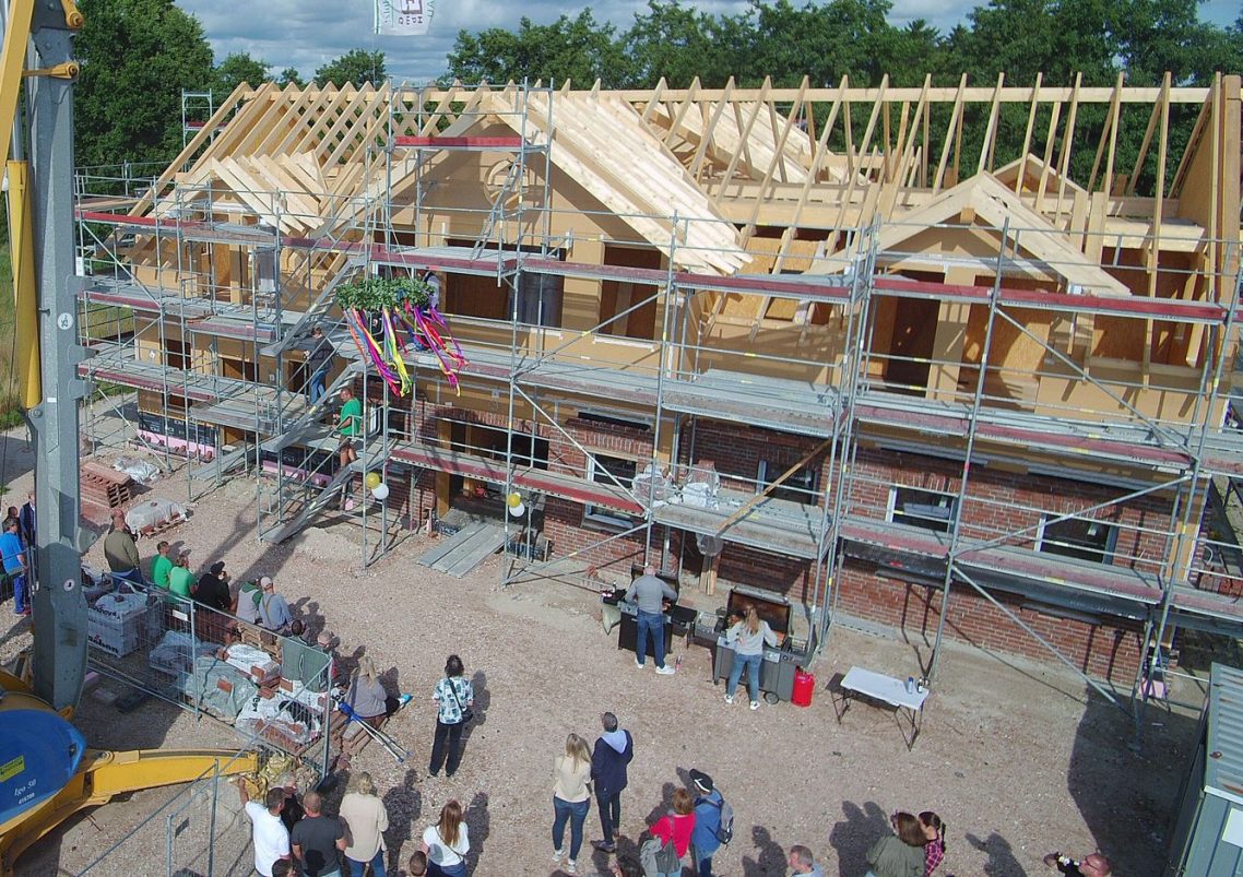 Ein großartiges Richtfest! Unser Dank gilt allen Handwerkern, allen Helfern und Gästen! Ein Rohbau eines Hauses mit Baugerüst, Menschen versammeln sich im Vordergrund.