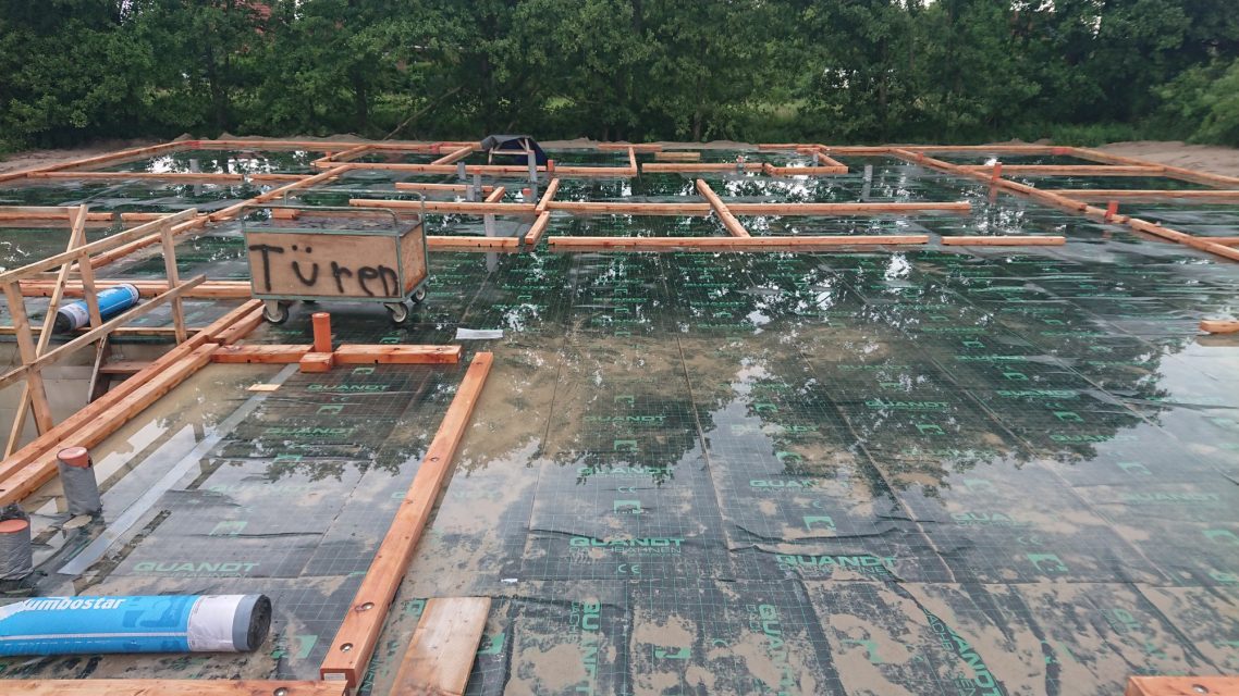 Bodenplatte muss austrocknen Baufläche mit Holzrahmen und grüner Folie, umgeben von Bäumen.