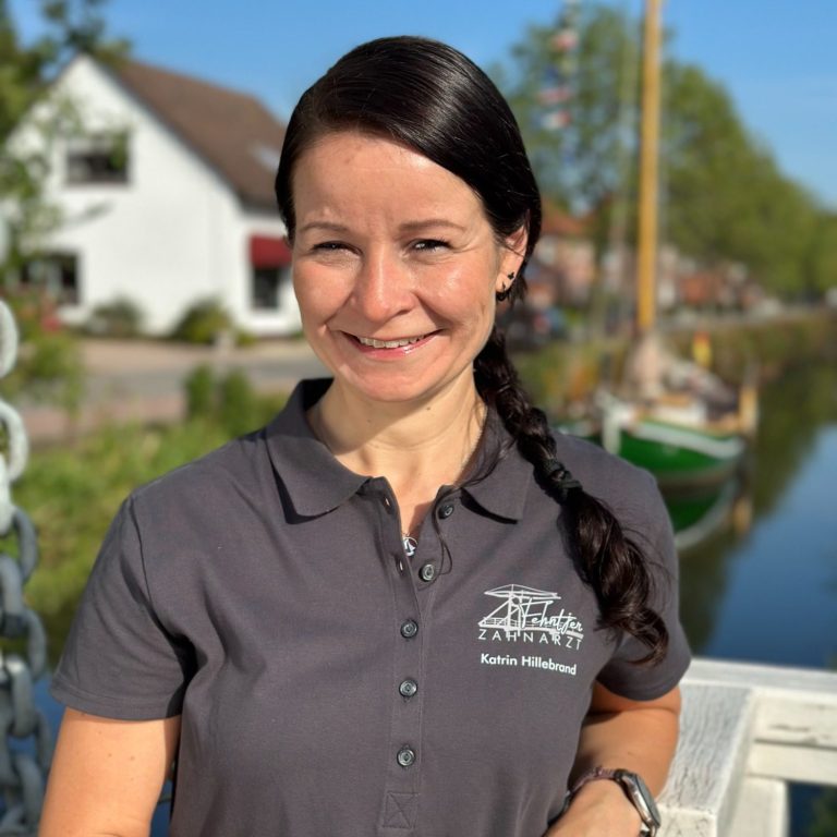 Frau mit langen, dunklen Haaren, trägt ein graues Shirt, steht lächelnd an einem Kanal mit Wasser.