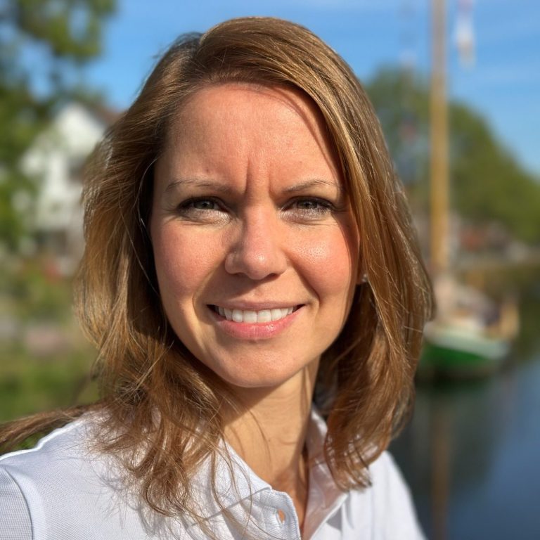Frau mit braunen Haaren lächelt vor einer natürlichen Kulisse am Wasser.