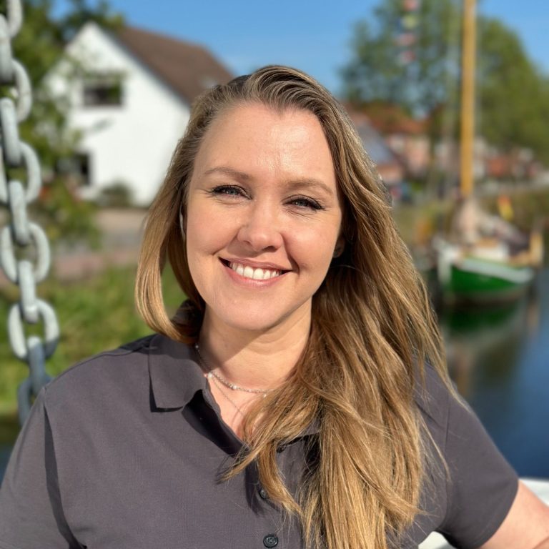 Lächelnde Frau mit langen Haaren steht am Wasser, im Hintergrund Boote und Gebäude.