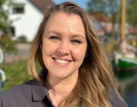 Lächelnde Frau mit langen, blonden Haaren vor einer malerischen Landschaft.