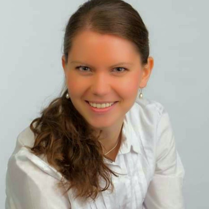 Porträt einer lächelnden Frau mit braunen Haaren, in einem weißen Hemd vor hellem Hintergrund.