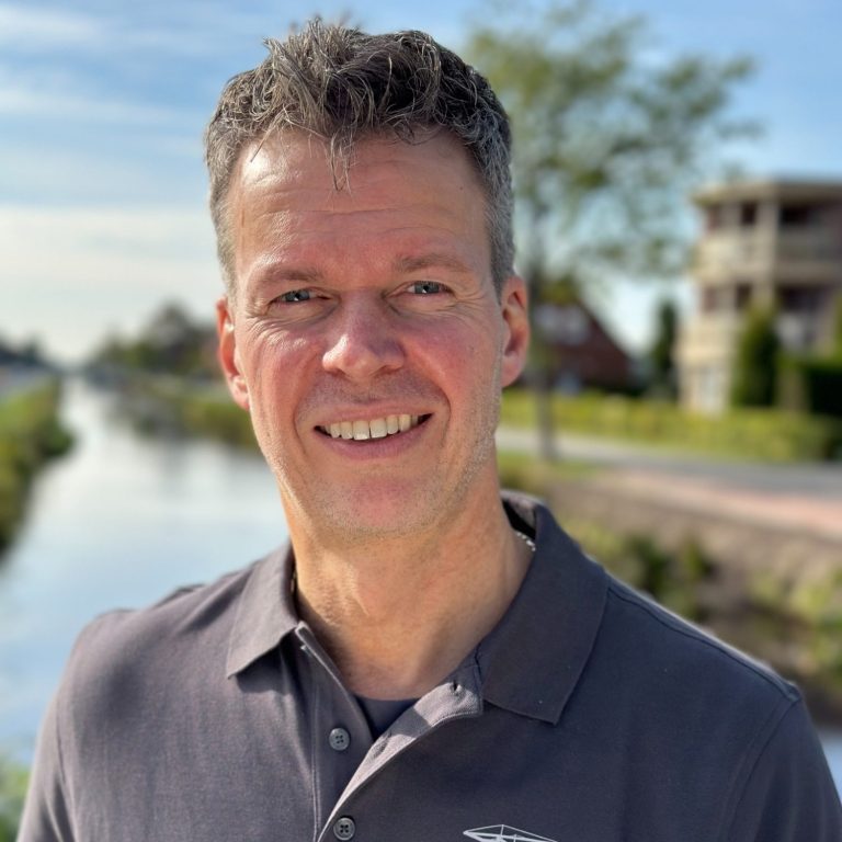 Lächelnder Mann in einem grauen Shirt vor einem Kanal und Wohngebäuden.