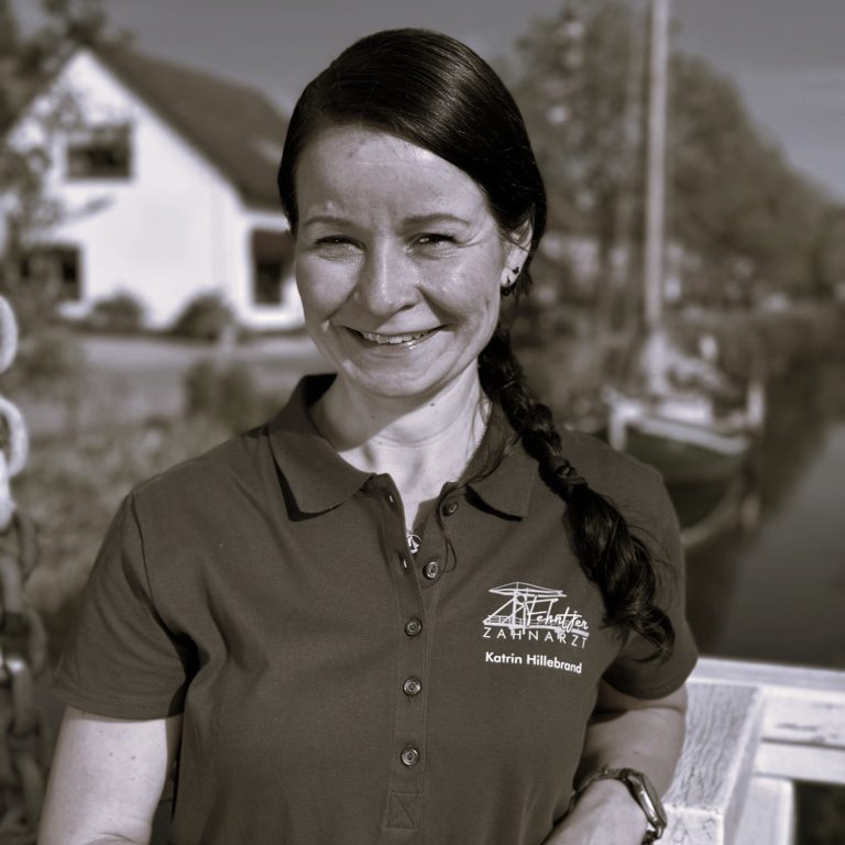 Frau mit langen, dunklen Haaren, trägt ein graues Shirt, steht lächelnd an einem Kanal mit Wasser.