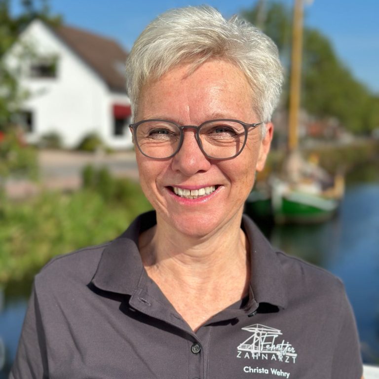 Lächelnde Frau mit blonden, kurzen Haaren, Brille und grauem Shirt am Wasser.
