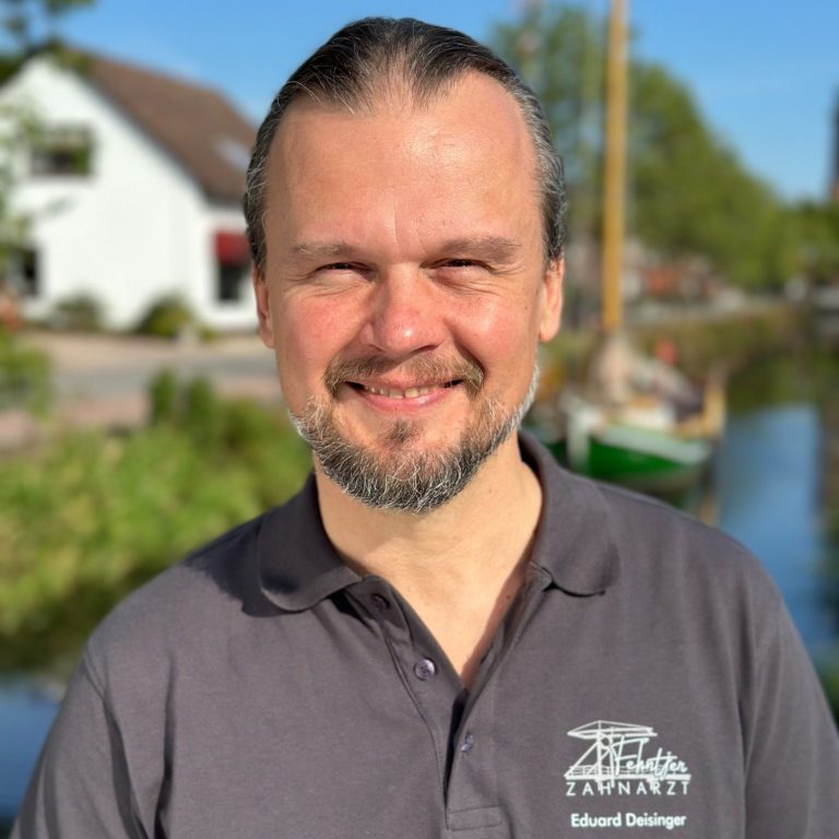 Mann mit langen Haaren, lächelnd, vor einer idyllischen Landschaft mit Wasser und Boot.